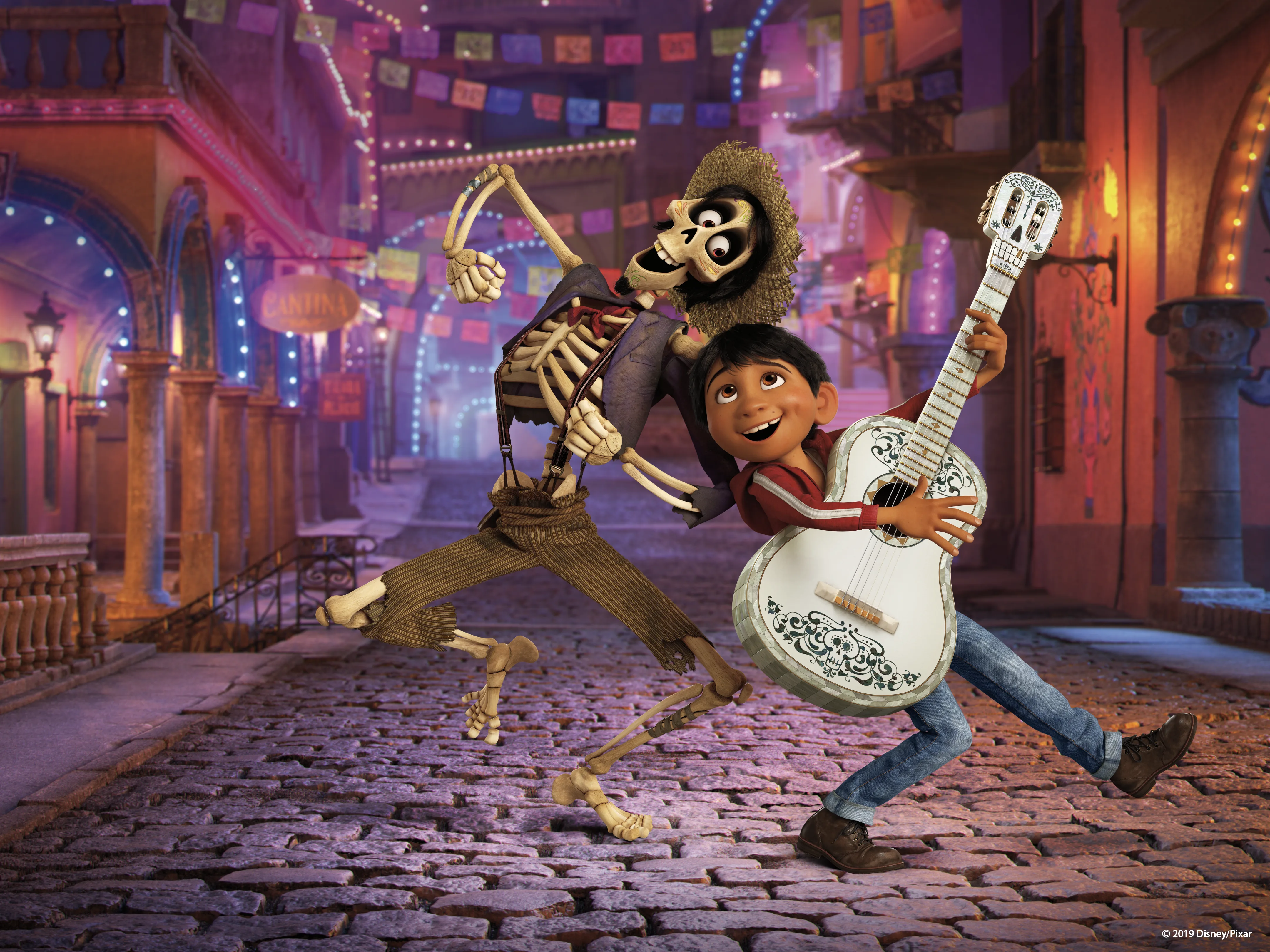 Héctor and Miguel pose with a white guitar beneath colorful papel picado banners on a street in the Land of the Dead in Disney/Pixar's Coco.