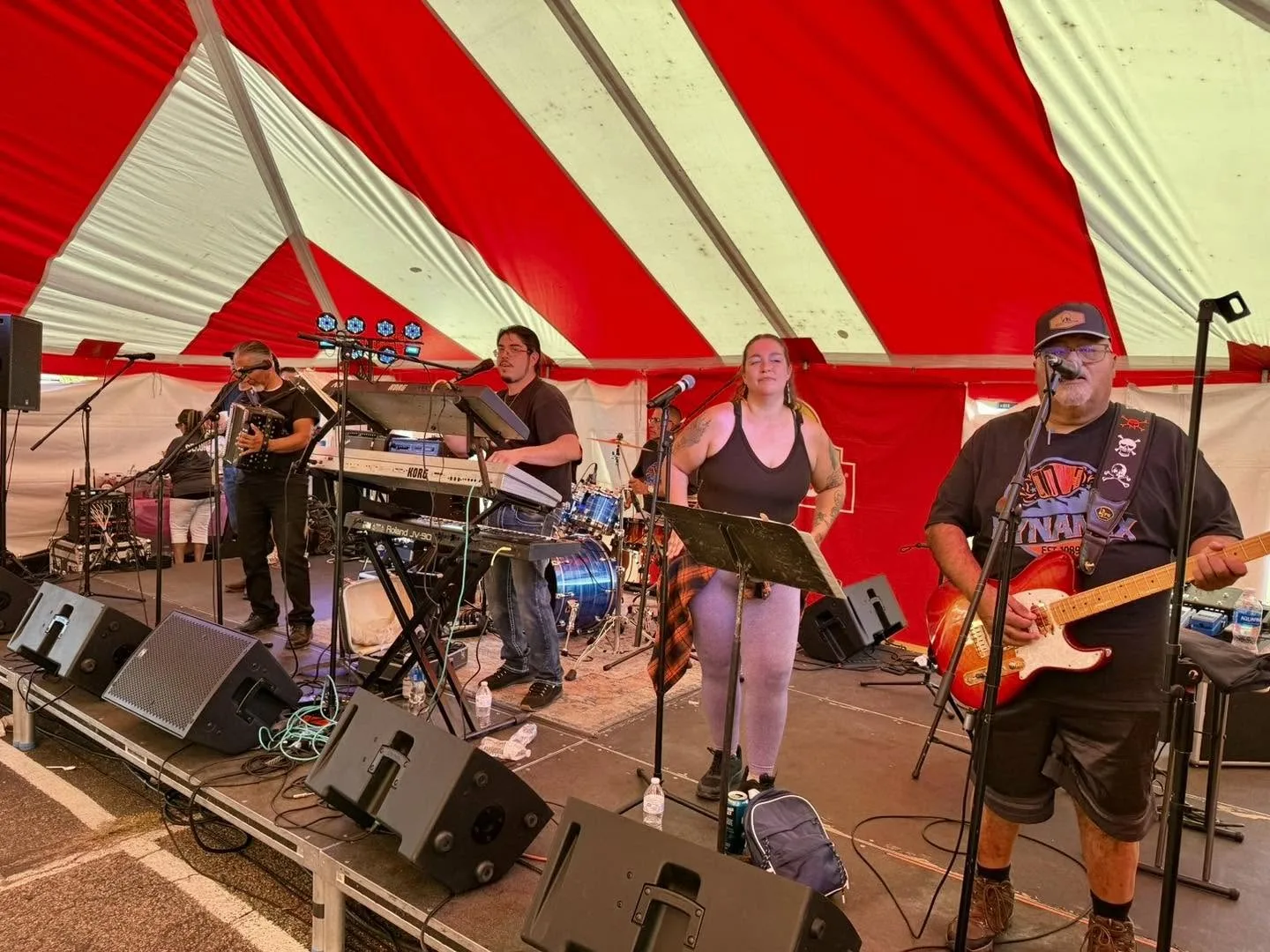 Dynamix performs with accordion, keyboards, vocals, drums, and guitar beneath a red-and-white tent.