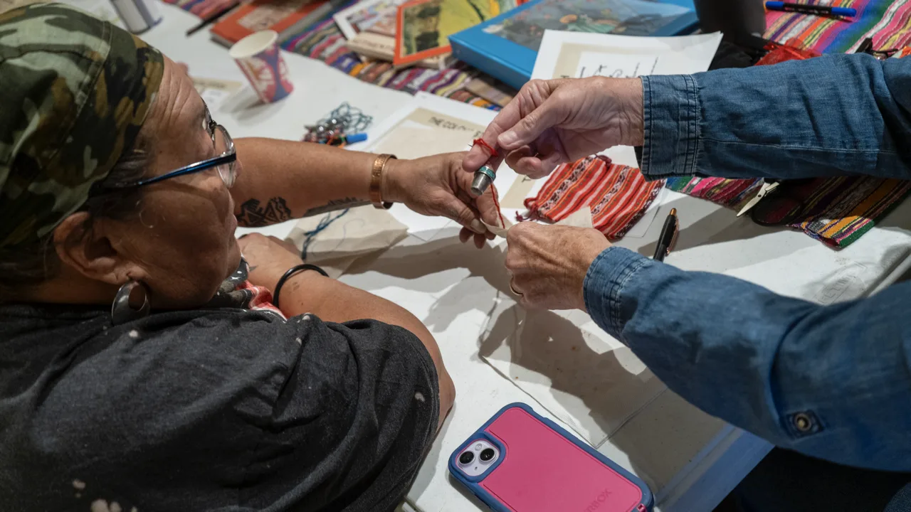 Two people work together with thread at a table covered with colorful art materials.