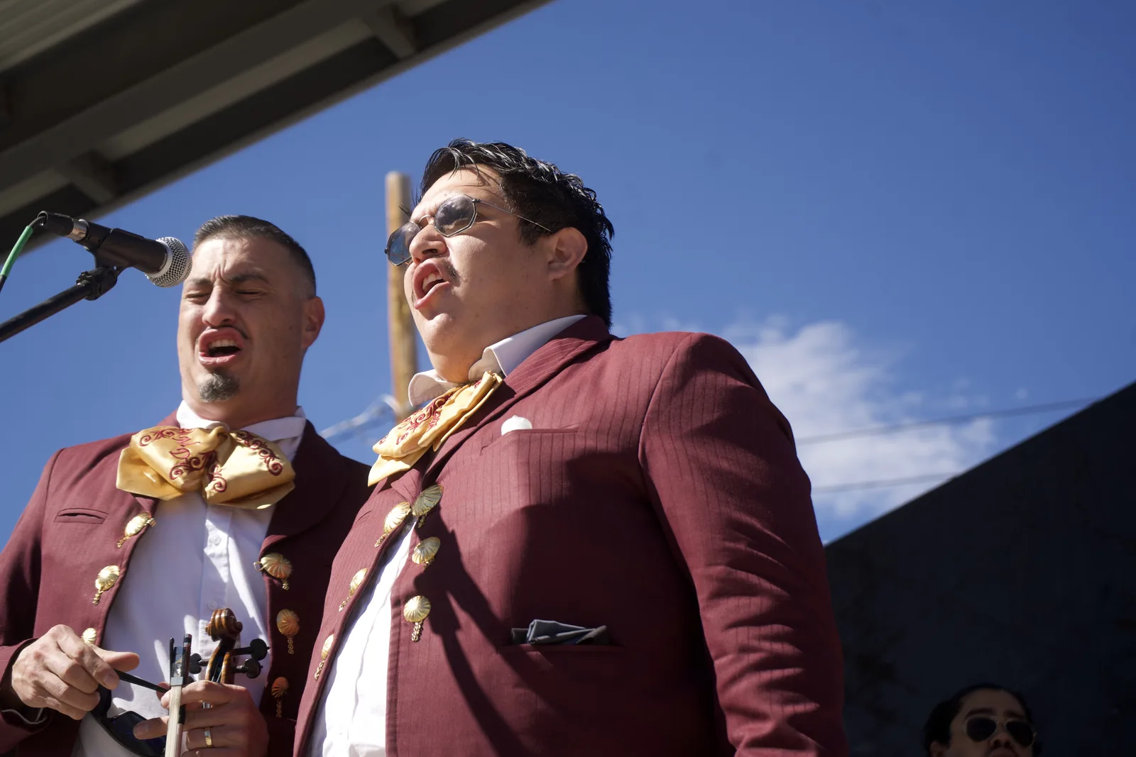 Two mariachi musicians sing together at an outdoor microphone.