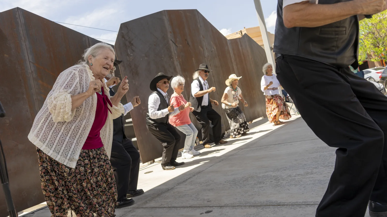Adults dance in a line outdoors beside sculptural steel panels.