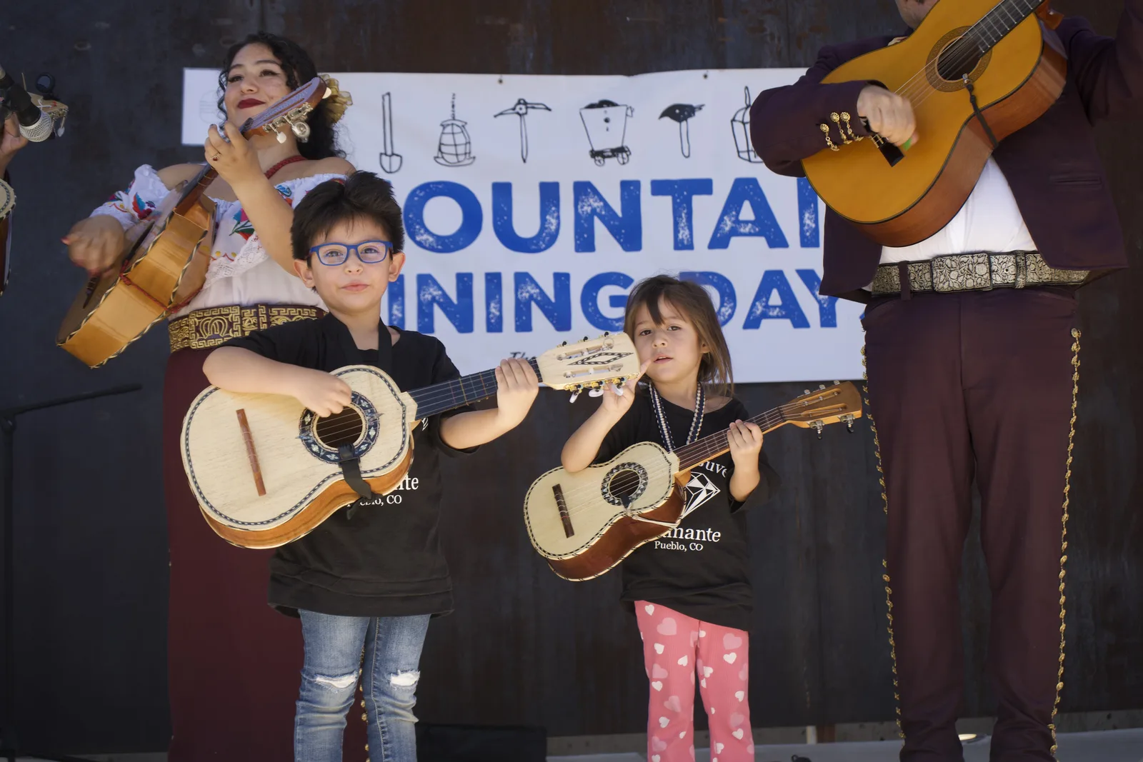 Two young musicians hold small string instruments beside mariachi performers.