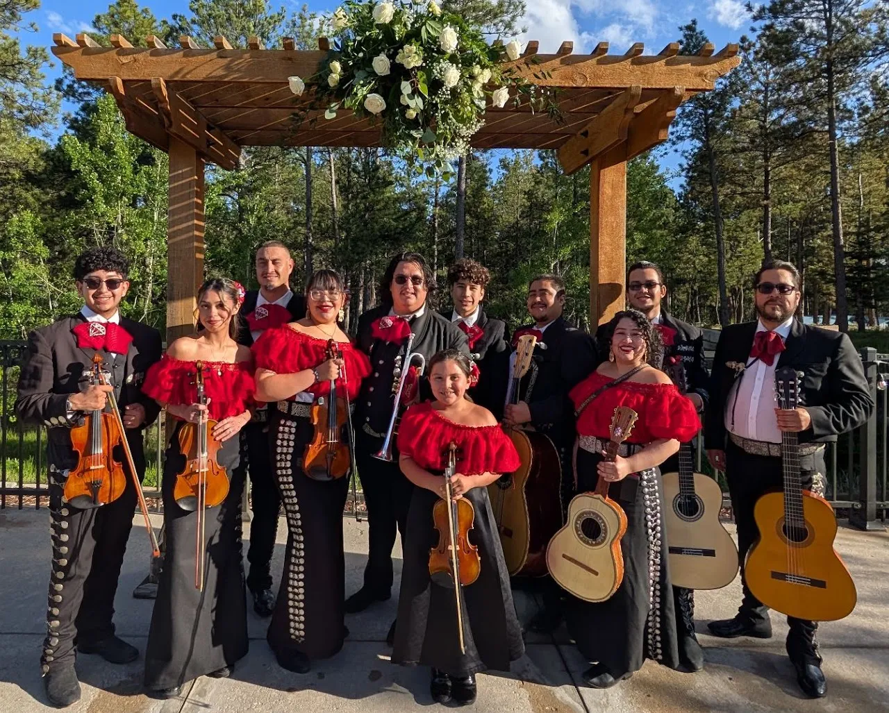 Mariachi Diamante members pose with violins, guitars, and brass instruments beneath a flower-covered pergola.