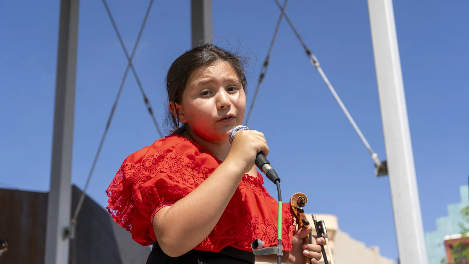 A young singer performs into a microphone outdoors in a red folklórico-style dress, with mariachi musicians behind her.