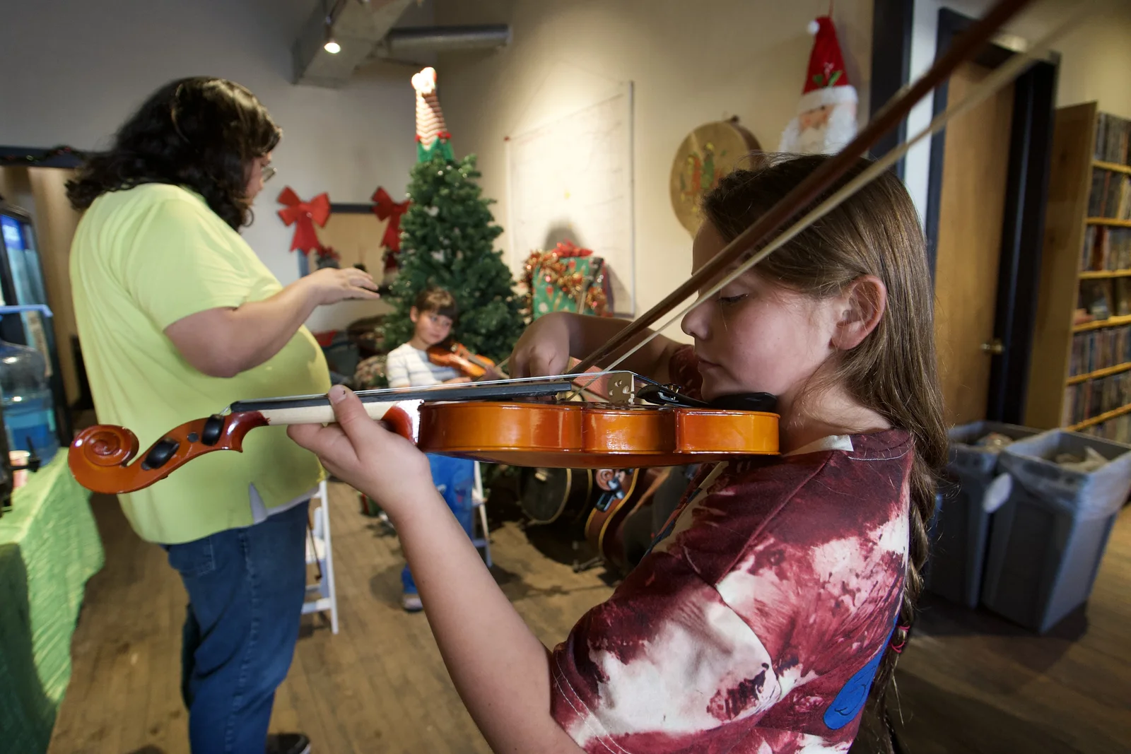 A young violinist practices with an instructor while another student plays in the background.