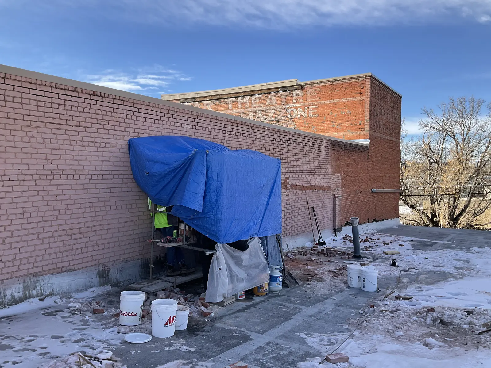 Blue temporary enclosure and masonry supplies beside the south brick wall in snow.